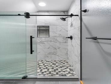 Angela & Dereck | Bathroom This bathroom remodel features natural wood cabinetry paired with a sleek white quartz vanity top and matte black fixtures. 