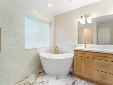 Vaie | Master Bathroom This master bathroom features marble-look porcelain floors, custom maple cabinetry, quartz countertops, and brushed gold fixture